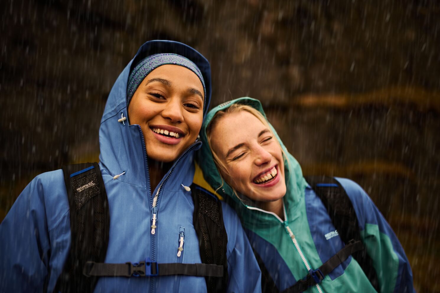 A woman leaning against another woman's shoulder whilst smiling, wearing waterproof jackets in the rain.